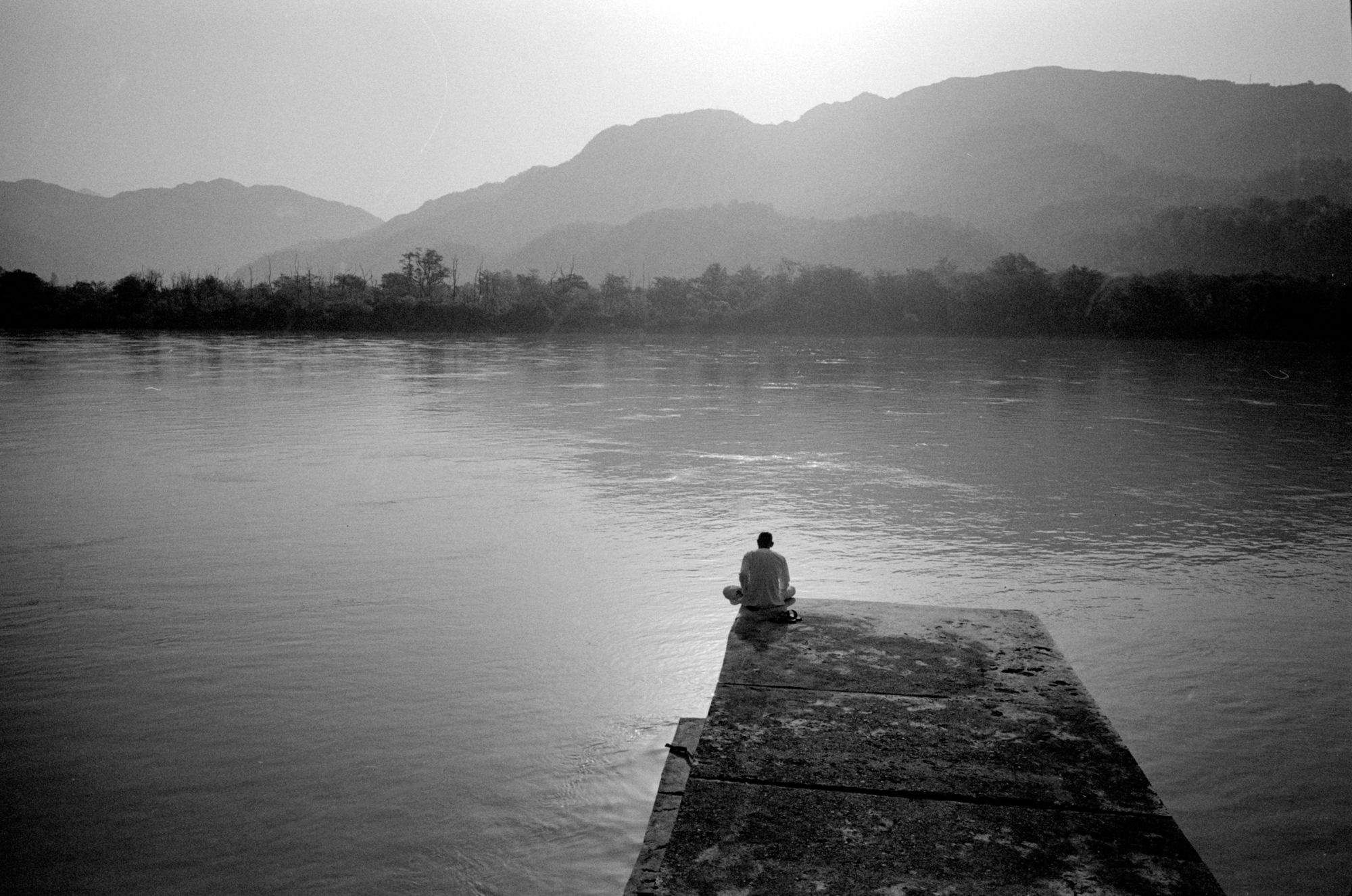 Man sitting alone on river pier, mountains in mist — India
